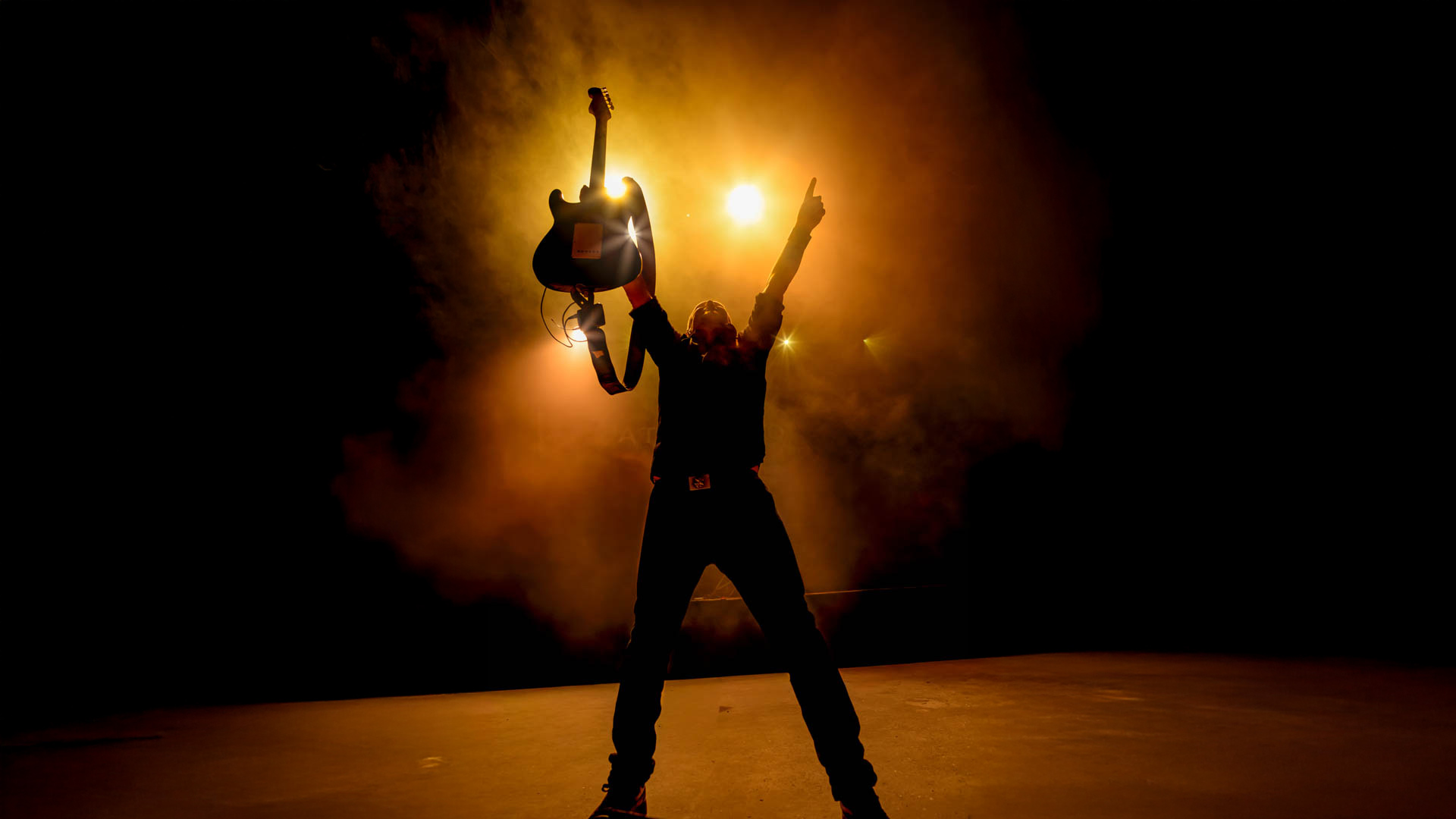 En mand står med hænderne i vejret på en scene. I den ene hånd har han en guitar, mens pegefingeren på den anden hånd peger op i luften. Der er røg omkring ham og et scenelys, der lyser direkte på ham.
